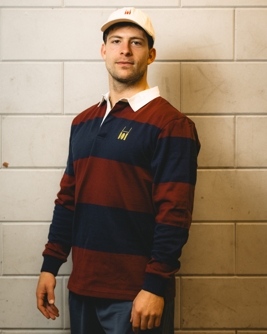 Male model in a flat cap wearing the Rugby Culture Navy & Maroon Rugby Polo against a white brick wall