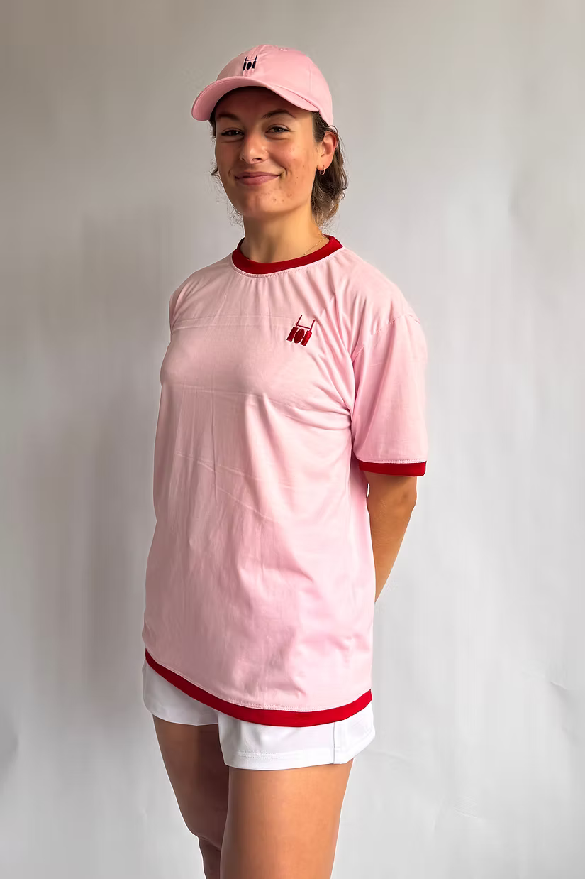 Rugby Culture Pink & Red Logo T-Shirt worn by a female model with a pink cap and white shorts, front view