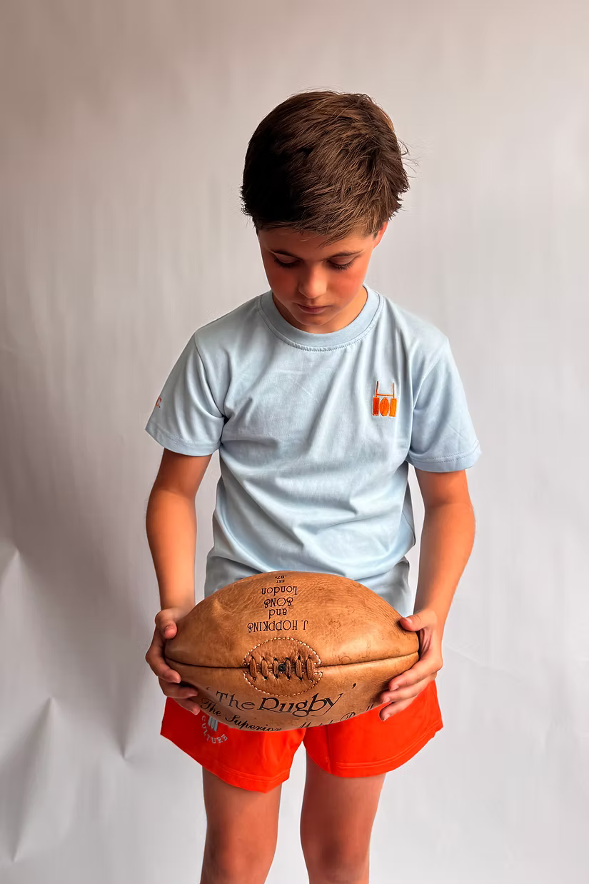Young rugby player in a sky blue Rugby Culture tee and orange kids' scrum shorts, holding a rugby ball