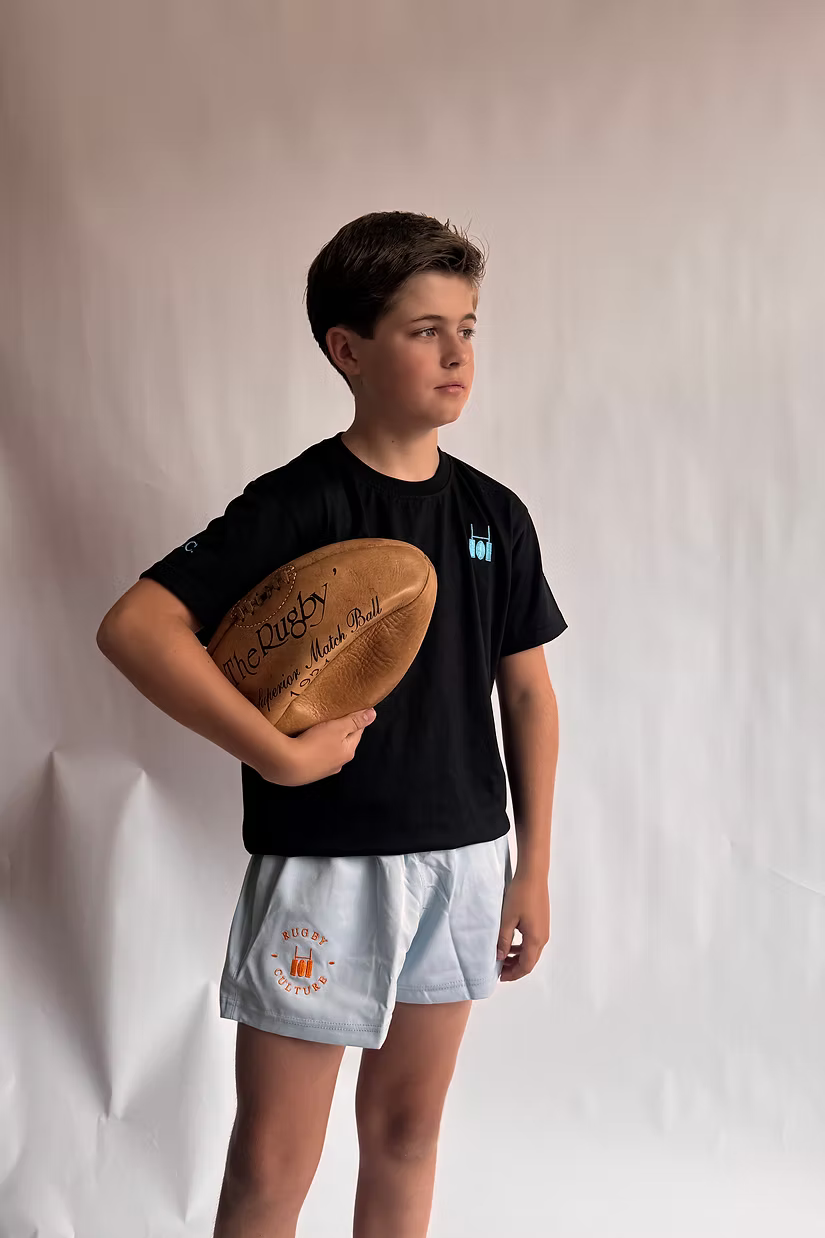 Rugby Culture Kids Baby Blue Scrum Shorts worn with a black tee by a young player holding a rugby ball