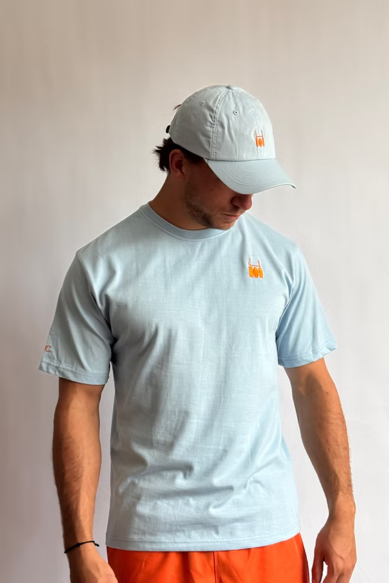 Rugby Culture Baby Blue Back Print T-Shirt worn by a male model with a white cap and orange shorts, front view