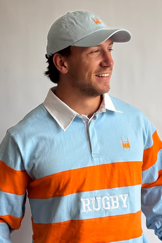 Male model in a cap wearing the Rugby Culture Sky Blue & Orange Rugby Polo with 'RUGBY' on the chest band